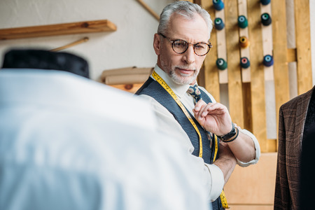 handsome senior tailor looking at jackets on mannequins at sewing workshopの写真素材