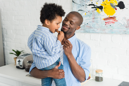 african american man holding little son with cookie in kitchen at homeの写真素材
