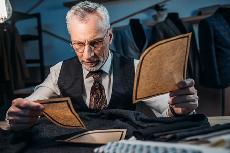 handsome mature tailor working with cut patterns at sewing workshop in late evening