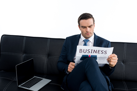 middle aged businessman reading newspaper while sitting on couch with laptop isolated on whiteの写真素材