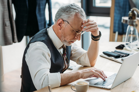serious mature tailor working with laptop at sewing workshopの写真素材