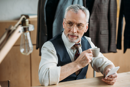 stylish mature tailor drinking coffee and using smartphone at sewing workshopの写真素材