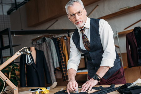 handsome mature tailor marking cloth pattern with chalk at sewing workshop and looking at cameraの写真素材