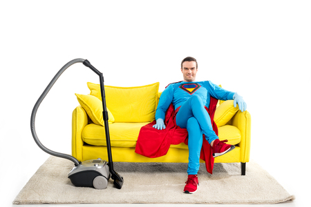 smiling male superhero in rubber gloves sitting on sofa near vacuum cleaner and looking at camera isolated on whiteの写真素材