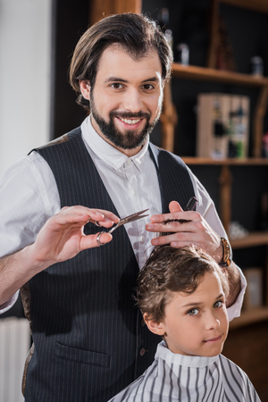 adorable little kid getting haircut from handsome smiling barber at kids barbershopの写真素材