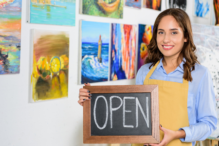 happy female artist holding signboard with word open in workshopの写真素材