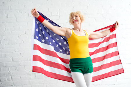 cheerful sporty senior woman in sportswear holding us flag and looking awayの写真素材
