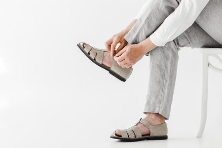 cropped image of male model in linen trousers sitting on chair and putting on sandals isolated on grey backgroundの写真素材