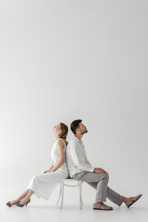 side view of young couple of models in linen clothes sitting back to back on chair isolated on grey backgroundの写真素材