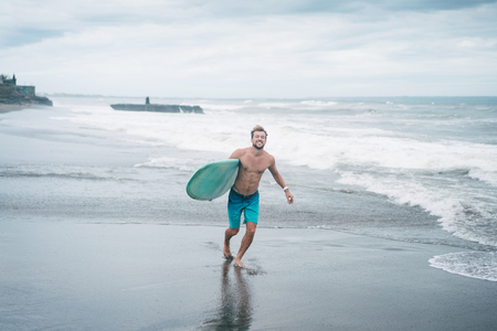 smiling surfer running with surfboard on beach in Bali, Indonesiaの写真素材