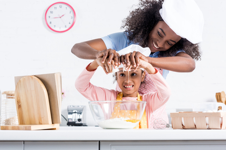 smiling african american mother and daughter in chef hats putting eggs into bowl together on kitchenの写真素材