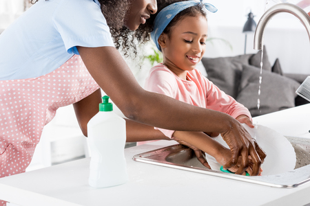 african american mother and daughter washing dishes with detergent on kitchenの写真素材