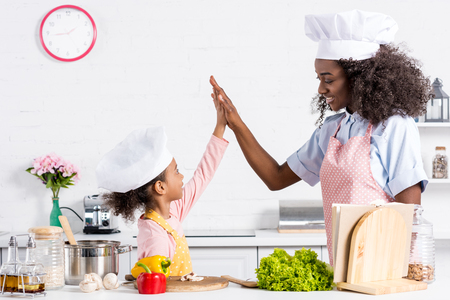 african american mother and daughter in chef hats cooking and giving high five on kitchenの写真素材