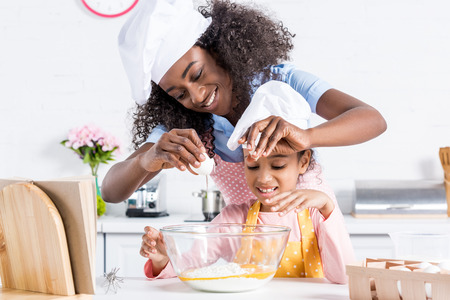 happy african american mother and daughter in chef hats making dough on kitchenの写真素材