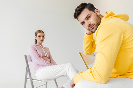 stylish young male and female models in pink and yellow hoodies sitting on chairs isolated on whiteの写真素材
