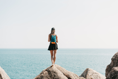 back view of female tourist with backpack standing on rocks and looking at the seaの写真素材