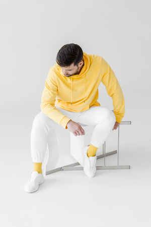 thoughtful young man in yellow hoodie sitting on flipped chair on whiteの写真素材