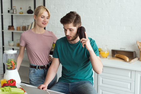 man pretending talking on eggplant at table with laptop and girlfriend near by in kitchen at home, vegan lifestyle conceptの写真素材
