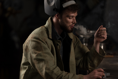 workman with lifted up protective mask sitting and smoking cigarette at factoryの写真素材