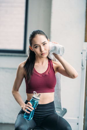 tired asian sportswoman with towel and water bottle after workout at gymの写真素材