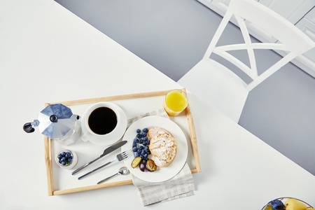 top view of croissant with fresh blueberries and plum pieces, glass of juice, cup of coffee and yogurt on wooden tray for breakfast on white tabletopの写真素材