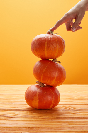 cropped shot of female hand touching stacked ripe pumpkins on wooden surface on orangeの写真素材
