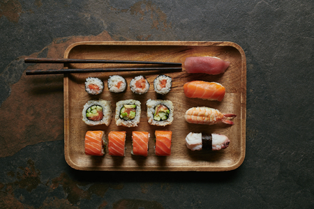 top view of arranged sushi set on wooden plate and chopsticks on dark surfaceの写真素材