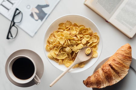 top view of bowl of dry cereal breakfast with cup of coffee and croissant on white surface with...