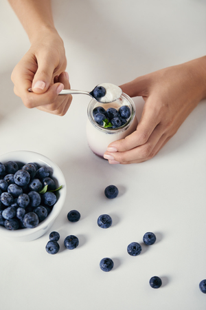 cropped shot of woman with yogurt and fresh blueberries for breakfast on white tabletopの写真素材
