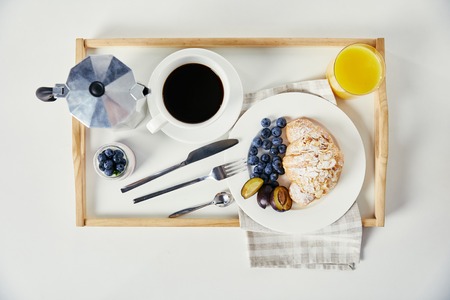 top view of croissant with fresh blueberries and plum pieces, glass of juice, cup of coffee and yogurt on wooden tray for breakfast on white tabletopの写真素材