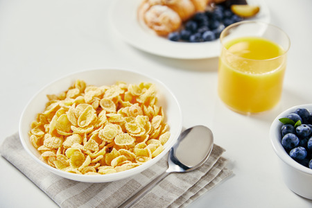 close up view of corn flakes in bowl, glass of juice and croissant with blueberries and plum pieces for breakfast on white tabletopの写真素材