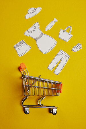 top view of little shopping trolley with paper clothes on yellow backdropの写真素材