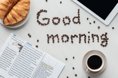 top view of cup of coffee and good morning lettering made of coffee beans on white surface with croissants, newspaper and tabletの写真素材