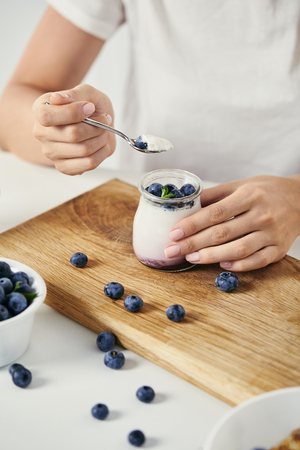 cropped shot of woman at tabletop with yogurt with fresh blueberries for breakfast on wooden cutting boardの写真素材