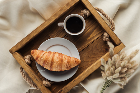 top view of coffee and croissant on tray with lagurus ovatus bouquet on beige cloth, breakfast in bed conceptの写真素材