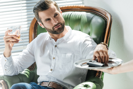 stylish man holding glass of whiskey while taking gun from tray