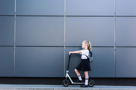 side view of adorable little schoolgirl with long curly hair riding scooter on street