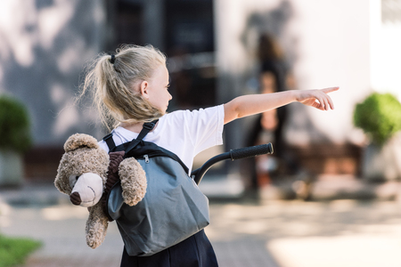 back view of adorable little schoolgirl with backpack and teddy bear riding scooter and pointing away with finger on streetの写真素材
