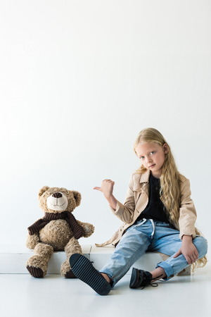 full length view of beautiful stylish kid pointing at teddy bear and looking at camera on whiteの写真素材