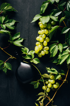 top view of fresh white grapes with green leaves and bottle of wine on dark wooden surfaceの写真素材