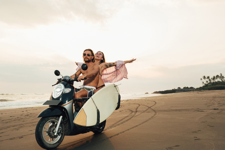happy boyfriend and girlfriend riding scooter with surfboard on beach in bali, indonesiaの写真素材