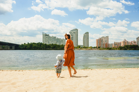 rear view of mother and son walking on sandy beach to river with city on backgroundの写真素材