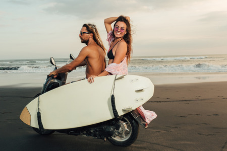 happy couple riding scooter with surfboard on beach in bali, indonesiaの写真素材