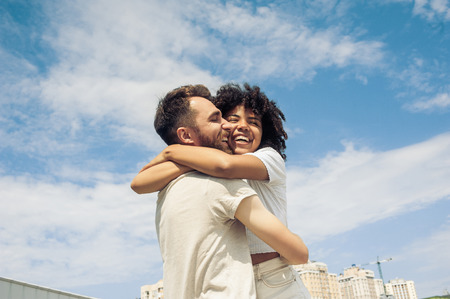 low angle view of happy young multiracial couple hugging against blue skyの写真素材
