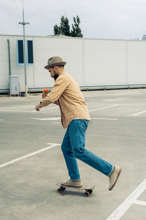 stylish young man holding disposable coffee cup and riding skateboard on streetの写真素材