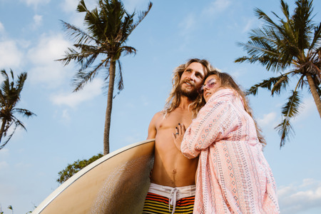 low angle view of girlfriend hugging smiling boyfriend with surfboard on beach in bali, indonesiaの写真素材