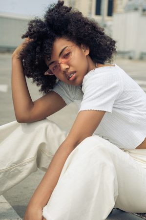 beautiful young african american woman in sunglasses sitting and looking at camera on streetの写真素材
