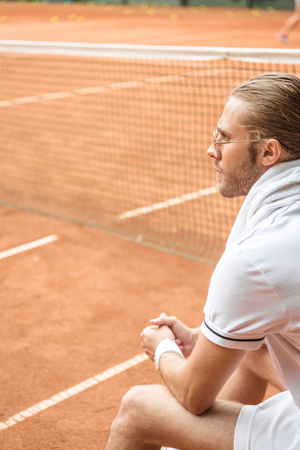 old-fashioned tennis player with towel resting on brown tennis court with netの写真素材