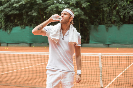 retro styled tired tennis player with towel drinking water on tennis courtの写真素材