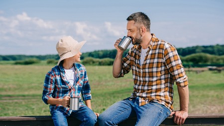 happy father and son sitting on fence and drinking from metal cups at farmの写真素材